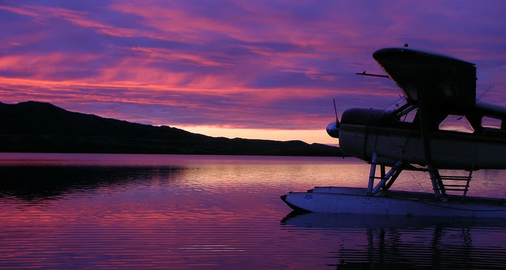Silhouette of a Newhalen Lodge floatplane docked at dusk with stunning purple and gold reflections